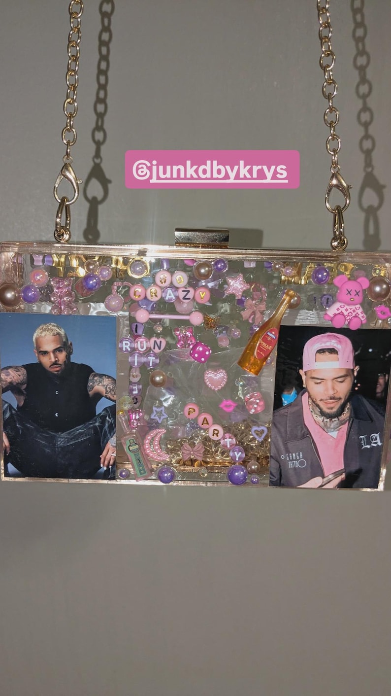 May include: Clear acrylic clutch with gold chain strap, filled with pink, purple, and clear embellishments. Features photos of a person, along with the words "RUN IT CRAZY PARTY". Includes a small champagne bottle charm.