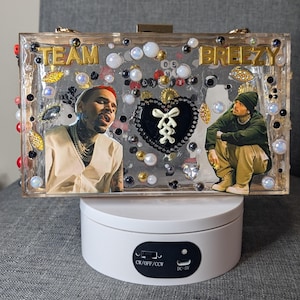 May include: A clear rectangular clutch with the words "TEAM BREEZY" in gold lettering. The clutch features images of a person, decorative beads, and a black heart-shaped design. The clutch is displayed on a white rotating stand.