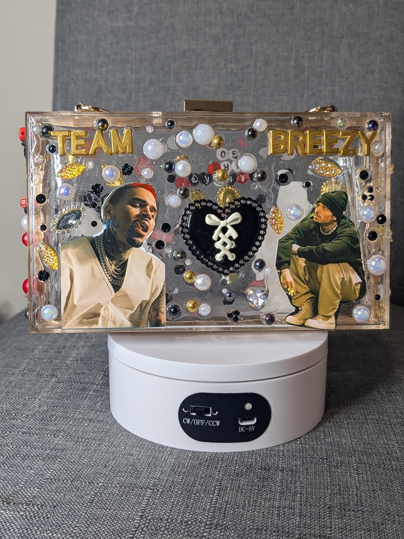 May include: A clear rectangular clutch with the words "TEAM BREEZY" in gold lettering. The clutch features images of a person, decorative beads, and a black heart-shaped design. The clutch is displayed on a white rotating stand.