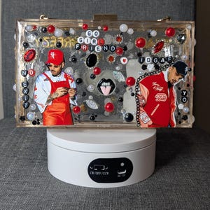 May include: A clear rectangular clutch decorated with red, black, and white embellishments, including beads and jewels. The clutch features images of a person and the words "GO GIRL FRIEND" and "IDGAF".