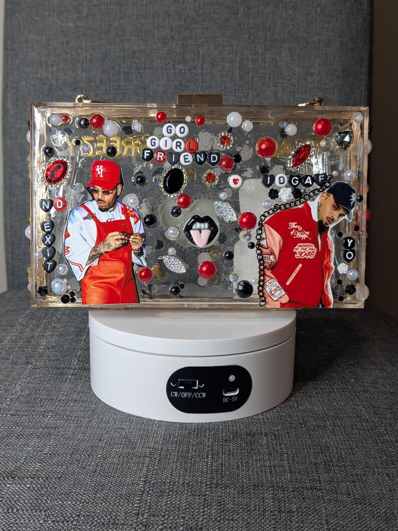 May include: A clear rectangular clutch decorated with red, black, and white embellishments, including beads and jewels. The clutch features images of a person and the words "GO GIRL FRIEND" and "IDGAF".
