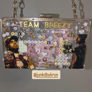 May include: A clear acrylic clutch with gold chain straps. The clutch is decorated with images, beads, and text that reads "TEAM BREEZY", "GIRLFRIEND", and "DON'T JUDGE ME". The clutch has a gold clasp and is filled with various decorative elements.