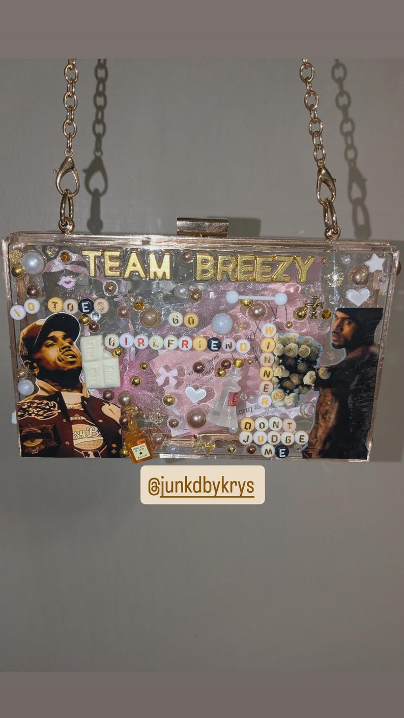May include: A clear acrylic clutch with gold chain straps. The clutch is decorated with images, beads, and text that reads "TEAM BREEZY", "GIRLFRIEND", and "DON'T JUDGE ME". The clutch has a gold clasp and is filled with various decorative elements.
