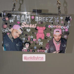 May include: Clear acrylic clutch with a gold chain strap, adorned with pink and black embellishments. The clutch features images of a person, the text "TEAM BREEZY Sweet Residuals", and the phrase "IT DEPENDS Gimme That".