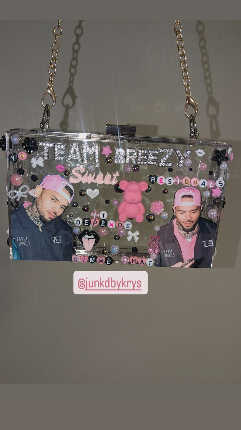 May include: Clear acrylic clutch with a gold chain strap, adorned with pink and black embellishments. The clutch features images of a person, the text "TEAM BREEZY Sweet Residuals", and the phrase "IT DEPENDS Gimme That".