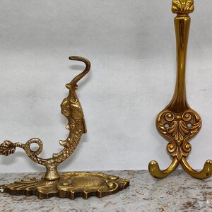 Puede incluir: Dos ganchos decorativos antiguos de latón. Uno presenta un diseño de pez y el otro tiene una parte superior decorativa con una base de tres puntas. Ambos ganchos son de color dorado y están sobre una superficie moteada.