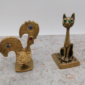 Può includere: Due statuette in ottone: un gallo con dettagli blu e rossi e un gatto con occhi verdi. Il gallo ha un corpo strutturato e un becco curvo. Il gatto è seduto su una base rettangolare.