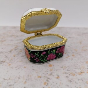 May include: Open, ornate jewelry box with a hinged lid. The box is black with pink and orange floral designs. The lid and trim are gold-colored, and the interior is white. The box is octagonal in shape.