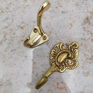 Könnte beinhalten: Zwei messingfarbene Metallhaken. Einer ist ein einfacher, gebogener Haken mit dreieckiger Basis und zwei Schraubenlöchern. Der andere ist ein verzierter Haken mit dekorativem Blumendesign und gebogenem Haken.