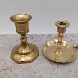 May include: Two brass candle holders. One is a simple, round design with a smooth base. The other has a round base with a decorative edge and a small handle.