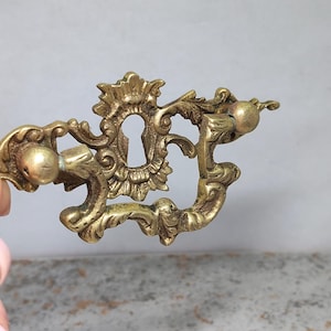 Pode incluir: Puxador de gaveta em latão dourado ornamentado com um buraco de fechadura e um design decorativo floral e de volutas. O puxador apresenta dois botões esféricos e é provavelmente para um móvel.