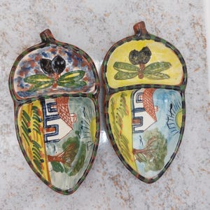Pode incluir: Dois pratos de cerâmica em forma de bolota com desenhos pintados à mão. Cada prato apresenta uma paisagem com uma casa, árvores e um sol, e uma secção com azeitonas e folhas. Os pratos têm um caule castanho e uma borda colorida.