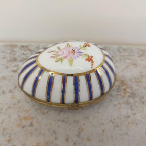 Op de afbeelding: Een witte porseleinen sieradendoos met een blauw en gouden gestreept ontwerp en een bloemenmotief op de deksel.