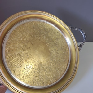 May include: A round serving tray featuring a gold-toned centre with an intricate embossed floral and scroll pattern. The tray has a smooth gold-toned rim and a silver-toned handle with detailed decorative metalwork on the side of the circular frame.