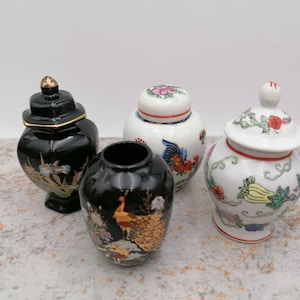 Puede incluir: Cuatro tarros decorativos de cerámica con tapas. Dos son negros con detalles dorados y diseños florales o de pavo real. Los otros dos son blancos con motivos florales y de aves coloridos. Los tarros varían en forma y tamaño.