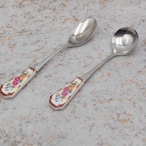 Pode incluir: Duas colheres de prata com cabos estampados com flores. Os cabos são brancos com uma borda vermelha e apresentam um design floral colorido. As colheres estão sobre uma superfície salpicada cinza e bege.