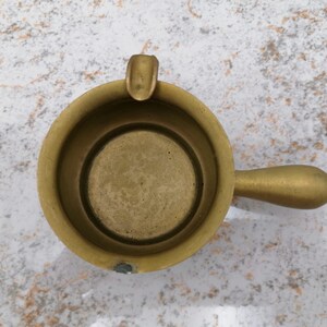 Pode incluir: Um cinzeiro de latão vintage com uma pequena alça e um descanso para cigarro embutido. O cinzeiro é circular com uma borda ligeiramente elevada e uma pequena alça arredondada. O latão tem uma pátina quente e envelhecida.