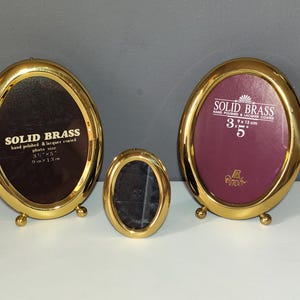 Op de afbeelding: Drie ovale fotolijsten van massief messing met een gepolijste afwerking en kleine bolvormige voetjes. De twee grotere lijsten hebben donkere inzetstukken met de tekst SOLID BRASS, hand polished & lacquer coated, 3.5 x 5 inches, 9 cm x 13 cm.