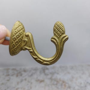 Op de afbeelding: Een goudkleurige, sierlijke haak met een ananasvormige bovenkant en een gebogen, decoratieve arm. De haak heeft een diamantpatroon basis en is ontworpen om items op te hangen. De haak is gemaakt van metaal en is waarschijnlijk voor decoratieve doeleinden.