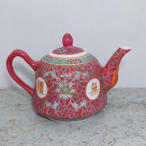 Pode incluir: Bule de cerâmica vermelho com tampa e alça. O bule é decorado com um padrão floral e caracteres chineses. O bico é branco. O bule é provavelmente usado para chá.