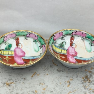 Peut inclure: Deux petits bols en porcelaine ornés de motifs peints à la main représentant un personnage en robe rose dans un jardin, avec des accents rouges, verts et roses. Chaque bol possède un bord doré et une base intérieure blanche.