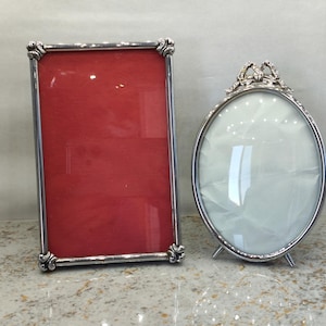 Pode incluir: Uma moldura retangular de prata com um inserto vermelho e uma moldura oval de prata com um inserto branco. A moldura retangular apresenta detalhes decorativos nos cantos, enquanto a moldura oval é rematada com um ornamento de prata em forma de laço.