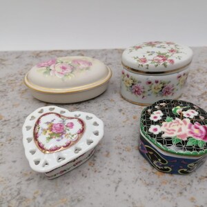Könnte beinhalten: Vier dekorative Schmuckdosen aus Porzellan. Eine ist oval mit cremefarbenem Grund und rosa Rosen. Eine weitere ist herzförmig mit durchbrochenem Design. Die anderen beiden sind rund, eine schwarz und die andere weiß, beide mit floralen Mustern.