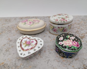 May include: Four decorative porcelain trinket boxes. One is oval with a cream base and pink rose design. Another is heart-shaped with a pierced design. The other two are round, one with a black base and the other with a white base, both with floral patterns.