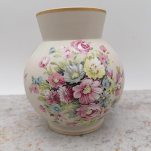 Op de afbeelding: Een crèmekleurige keramische vaas met een ronde body en een uitlopende hals. De vaas is versierd met een bloemmotief met roze, witte en gele bloemen met groene bladeren. De rand en de basis hebben een gouden rand.