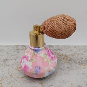 Puede incluir: Un atomizador de perfume vintage. La botella de cerámica está decorada con flores rosas, blancas y moradas y hojas verdes. Tiene una tapa de metal dorado y una pera tejida marrón. Un objeto decorativo.