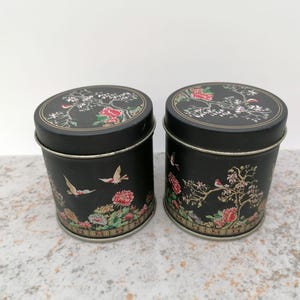 Pode incluir: Duas latas de metal preto com tampas, com desenhos florais e de pássaros. As latas cilíndricas têm bordas prateadas. As tampas e os lados são adornados com flores coloridas e pássaros voando.