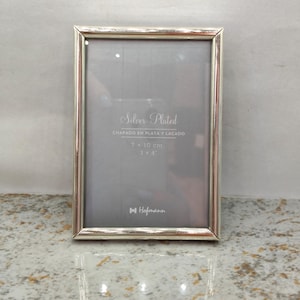 Pode incluir: Uma moldura retangular banhada a prata com acabamento metálico. O encarte exibe o texto Silver Plated, CHAPADO EN PLATA Y LACADO, 7 x 10 cm, 3 x 4 inches e o logotipo da marca Hofmann. A moldura tem um design de borda simples.