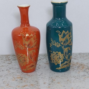 Pode incluir: Dois vasos decorativos. Um é laranja com desenhos florais dourados, o outro é verde-azulado com desenhos florais dourados. Ambos os vasos têm um pescoço estreito e uma base mais larga. Os vasos são provavelmente feitos de cerâmica ou porcelana.