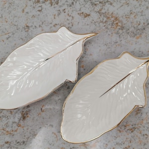 May include: Two white, leaf-shaped ceramic dishes with gold trim. The dishes have a detailed feather design and are set against a mottled grey background. The gold trim accents the edges of the leaf-shaped dishes.