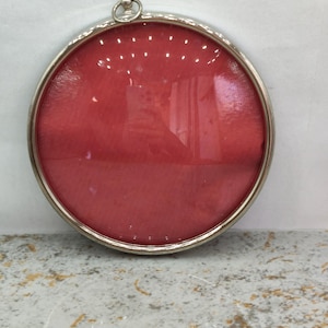 Pode incluir: Uma moldura de parede circular com uma borda metálica em tom prateado e uma frente de vidro convexo. O interior é preenchido com um tecido vermelho vibrante e sólido. Um pequeno anel metálico circular está fixado no topo da moldura.