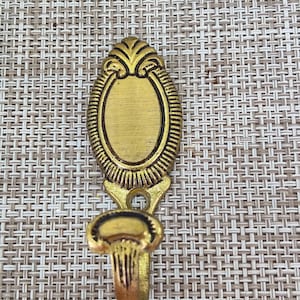 Könnte beinhalten: Ein goldfarbener Metall-Wandhaken mit einem ovalen Oberteil und einem dekorativen Haken. Die Oberseite hat ein verziertes Design mit einer leeren ovalen Mitte. Der Haken ist zum Aufhängen von Gegenständen konzipiert und eignet sich für verschiedene Einrichtungsstile.