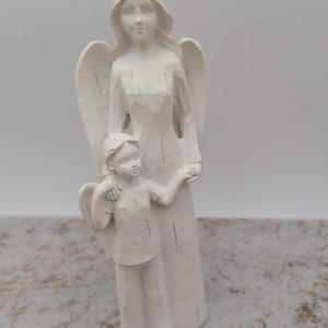 Pode incluir: Uma estatueta de anjo branco segurando uma criança. O anjo tem asas grandes e está vestindo um vestido longo. A criança tem asas pequenas e está vestindo uma camisa e calças. A estatueta é feita de um material liso.