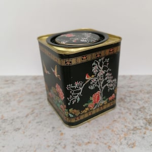 May include: Black square tin with a lid, featuring a floral and bird design. The tin has gold trim and a decorative border. The lid has a floral design. The tin is likely for storage or display.