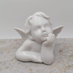 Pode incluir: Uma figura de querubim de cerâmica branca, apoiada no braço com uma expressão pensativa. O querubim tem pequenas asas e cabelo encaracolado. A figura é exibida em uma superfície clara e salpicada.