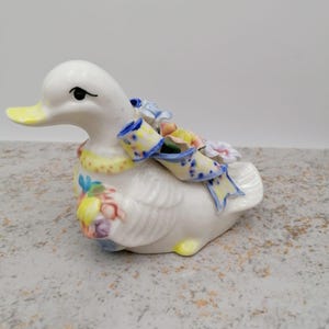 Pode incluir: Uma figura de pato de cerâmica branca com bico e detalhes amarelos. O pato tem uma fita estampada azul e amarela e é adornado com flores coloridas. Um item decorativo.