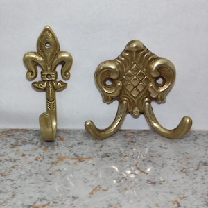 May include: Two brass wall hooks with metallic gold finishes. The left hook features a fleur-de-lis design with a single upward-curving prong. The right hook has a wider, ornate base with a diamond-patterned center and two outward-curving prongs.
