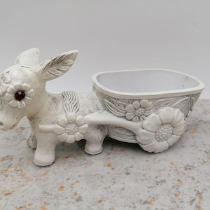 Puede incluir: Una maceta de burro blanco con un carro, adornada con detalles florales. El burro tiene ojos rojos y tira de una jardinera en forma de carro. La jardinera está decorada con flores y hojas. El diseño es caprichoso y decorativo.