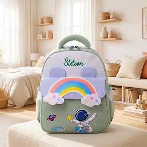 Op de afbeelding: Een saliegroene rugzak met een wit bovenpaneel waarop de naam Stetson in het groen is geborduurd. De tas heeft een lavendelkleurige flap met een regenboog- en wolkenafbeelding, twee kleine lavendelkleurige oren en een onderste gedeelte met een astronaut op een skateboard, een planeet, een gele ster en de tekst City Tour.