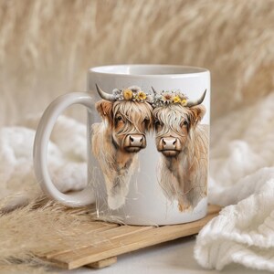 May include: White ceramic mug with a colorful floral design featuring two Highland cows with long, fluffy hair. The cows are wearing flower crowns.