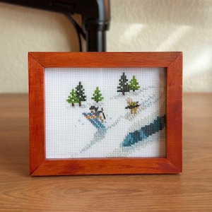 May include: A framed cross-stitch depicting a winter scene with green trees and a skier on a snowy slope. The frame is made of reddish-brown wood, and the artwork is displayed on a wooden surface.