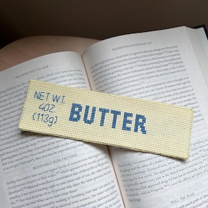Op de afbeelding: Een geborduurd label met het woord "BUTTER" in blauw op een crèmekleurige achtergrond. Het label toont "NET WT. 4OZ. (113g)". Het label ligt op een open boek met tekst over haaruitval.
