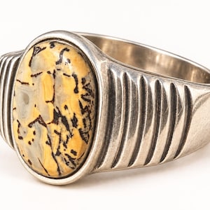 Op de afbeelding: Een zilveren ring met een grote, ovaalvormige steen met een geel, crème en grijs marmerpatroon, geaccentueerd door donkere, vertakte, aderachtige insluitingen. De metalen band heeft een gepolijste afwerking met verticale groeven.
