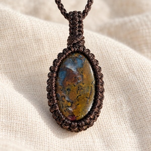 May include: An oval-shaped pendant featuring a multi-colored stone with brown, blue, and yellow tones, encased in a dark brown macrame cord setting. The pendant hangs from a matching dark brown braided cord necklace.