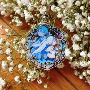 Puede incluir: Un charm de acrílico transparente con dos personajes de estilo anime en tonos azules y blancos. Uno tiene cabello azul y alas, el otro tiene cabello blanco y un bastón. El charm está sujeto a un cierre plateado.