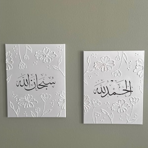 Peut inclure: Deux œuvres d'art sur toile blanche avec des motifs floraux en relief et de la calligraphie arabe noire. Les œuvres présentent les phrases « Subhan Allah » et « Alhamdulillah ». Les toiles sont rectangulaires et montées sur un mur vert clair.